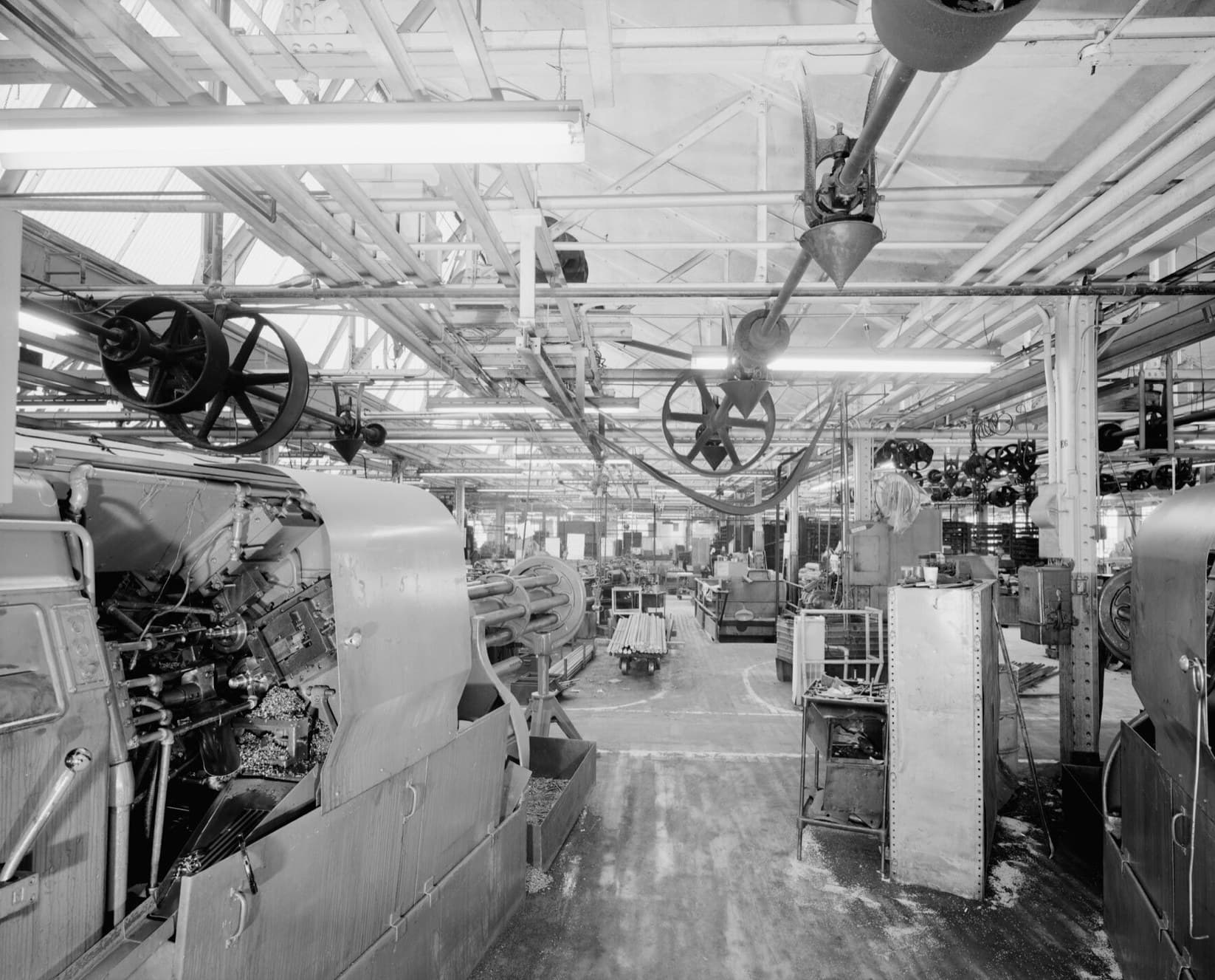 A long machine shop aisle with the original line shafting and belt wheels still hanging from the ceiling above rows of machines.