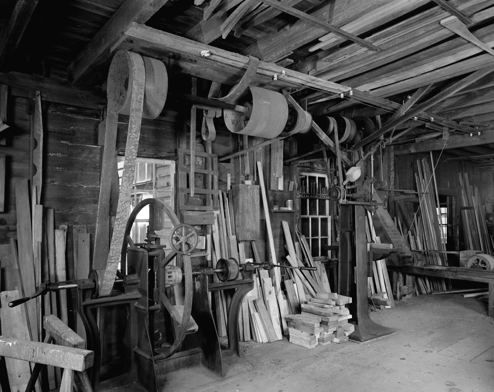 A timber wood shop where wide belts drop from overhead pulleys to a boring machine, a mortiser and a sander.